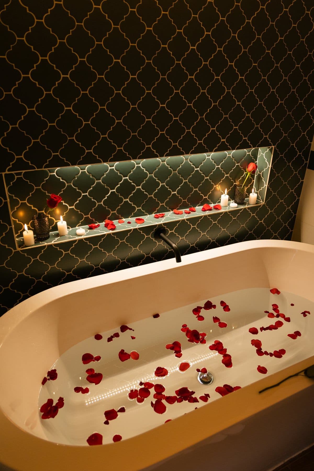 Bathtub scattered with red rose petals beside a candlelit niche