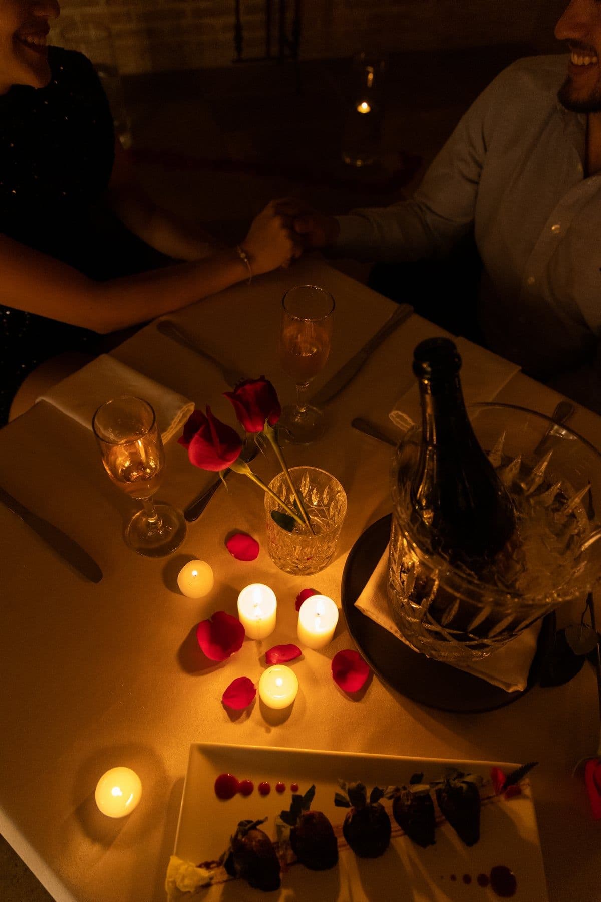 Couple holding hands over a candlelit table with roses, sparkling wine and chocolate-covered strawberries
